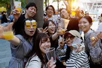 A group of cheerful young adults hold up drinks and smile at an outdoor gathering. One person wears novelty glasses with attached beer mugs. The scene is lively and festive, with sunlight shining in the background.
