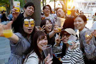A group of cheerful young adults hold up drinks and smile at an outdoor gathering. One person wears novelty glasses with attached beer mugs. The scene is lively and festive, with sunlight shining in the background.