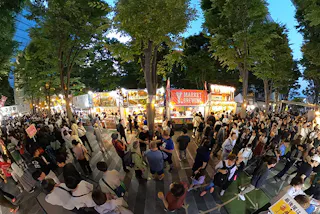 A large crowd gathers at an outdoor market festival in the evening, surrounded by tall trees and brightly lit food stalls. People are standing, walking, and queuing at various vendor booths under a clear sky.