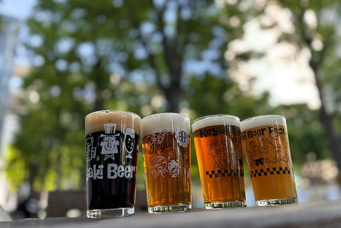 Four glasses of beer, each filled with different types and colors of beer, sit on a wooden surface outdoors. Green trees and a blurred cityscape can be seen in the background.
