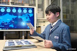A boy in a blue suit sits at a desk, looking at photographs from an album. Behind him is a world map on a screen with clocks showing times in different cities. The setting appears to be an educational or office environment.