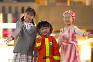Three smiling children stand together, each dressed in different costumes: one in a suit, one in a firefighter uniform, and one in a red-and-white striped apron and headscarf, posing indoors with outstretched arms.