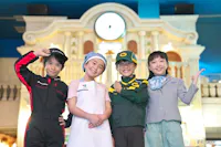 Four smiling children stand together, each dressed in different professional uniforms: a race car driver, a nurse, a delivery worker, and a flight attendant, posing in front of a building with a clock.