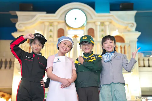 Four smiling children stand together, each dressed in different professional uniforms: a race car driver, a nurse, a delivery worker, and a flight attendant, posing in front of a building with a clock.
