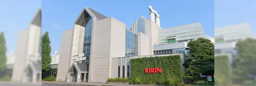 A modern, multi-story building with glass and white paneling, greenery on one wall, and a red "KIRIN" sign. Tall chimneys and trees surround the structure under a clear blue sky.