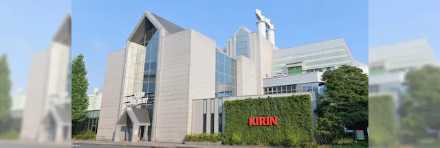 A modern, multi-story building with glass and white paneling, greenery on one wall, and a red "KIRIN" sign. Tall chimneys and trees surround the structure under a clear blue sky.