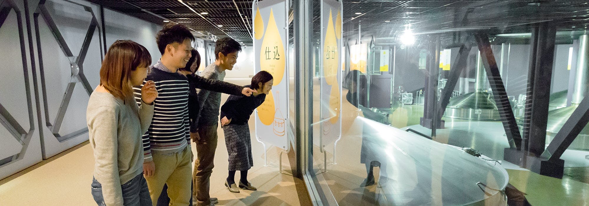 Four people stand in a corridor, looking and pointing through a large window at an exhibit or machinery in a brightly lit, industrial-looking space on the other side of the glass.