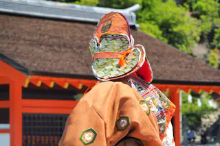 A person wearing a traditional, elaborate Japanese costume and headpiece stands outdoors in front of a building with orange wooden beams and a thatched roof. Lush greenery is visible in the background.