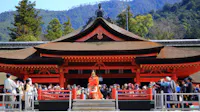 A performer in traditional costume stands in front of a large, ornate red Japanese shrine with a curved roof, as a crowd of people watches and takes photos. Trees and mountains are visible in the background.