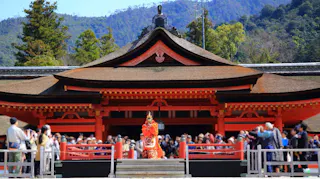 A performer in traditional costume stands in front of a large, ornate red Japanese shrine with a curved roof, as a crowd of people watches and takes photos. Trees and mountains are visible in the background.