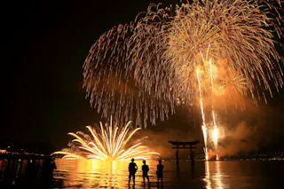 Silhouettes of people stand by the water, watching large golden fireworks explode in the night sky above a traditional torii gate, with reflections shimmering on the surface.