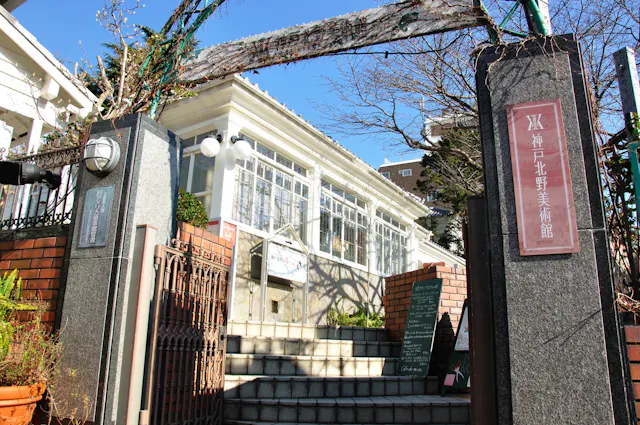 A stone and brick entrance with signs in Japanese leads up steps to a white, glass-enclosed building under a clear blue sky. There are leafless trees and a chalkboard with writing near the doorway.