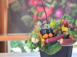 A black container holds an arrangement of colorful vegetables, including okra, cherry tomatoes, squash, greens, and herbs, set on a light wooden surface with a blurred, vibrant background.