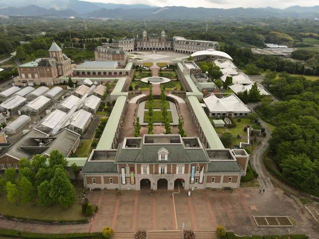 Aerial view of a large, elegant building complex with green roofs, gardens, greenhouses, paths, and surrounding trees, set against a backdrop of rolling hills and a cloudy sky.