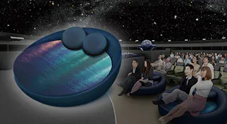 A planetarium with people seated and watching a starry night projection. In the foreground, a large round cushioned seat with two pillows is highlighted, contrasting with the regular seats.