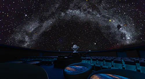 A dark planetarium with rows of empty seats and a star projector in the center, displaying a realistic, star-filled night sky with the Milky Way across the domed ceiling.
