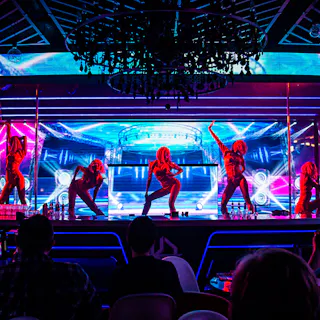 Kujira Entertainment Six dancers perform on a brightly lit stage with vivid neon blue and pink lights, while an audience watches from darkened tables in the foreground. A chandelier hangs above the scene.