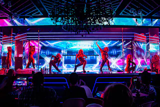 Six dancers perform on a brightly lit stage with vivid neon blue and pink lights, while an audience watches from darkened tables in the foreground. A chandelier hangs above the scene.