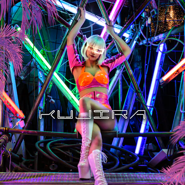 Kujira Entertainment A person in a shiny pink and orange outfit with white boots and a platinum bob hairstyle poses on a metal structure surrounded by neon lights and pink artificial palm leaves. The word "KUJIRA" appears across the image.