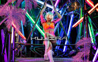 A person in a shiny pink and orange outfit with white boots and a platinum bob hairstyle poses on a metal structure surrounded by neon lights and pink artificial palm leaves. The word "KUJIRA" appears across the image.