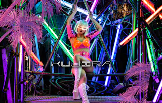 A person in a shiny pink and orange outfit with white boots and a platinum bob hairstyle poses on a metal structure surrounded by neon lights and pink artificial palm leaves. The word "KUJIRA" appears across the image.
