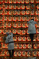 Two people in traditional clothing adjust rows of illuminated red paper lanterns with bold black kanji characters, creating a vibrant display at a festival or cultural event.