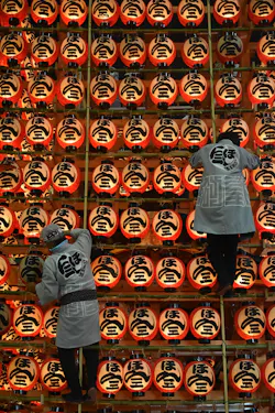 Two people in traditional clothing adjust rows of illuminated red paper lanterns with bold black kanji characters, creating a vibrant display at a festival or cultural event.