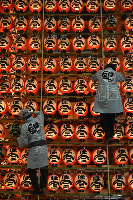 Two people in traditional clothing adjust rows of illuminated red paper lanterns with bold black kanji characters, creating a vibrant display at a festival or cultural event.