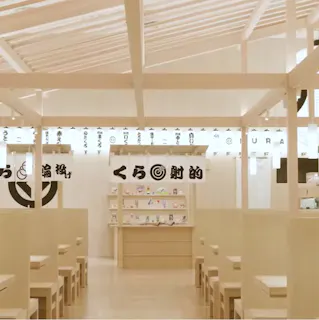 Modern Japanese restaurant interior with light wooden tables and benches, white ceiling beams, lantern-style lights, and shelves displaying small items. Japanese characters are visible on banners and lanterns above.