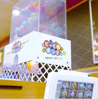 A colorful capsule toy dispenser with Japanese text, releasing a yellow capsule. Capsules are visible inside the machine, and a touchscreen menu displaying sushi items is in the foreground.