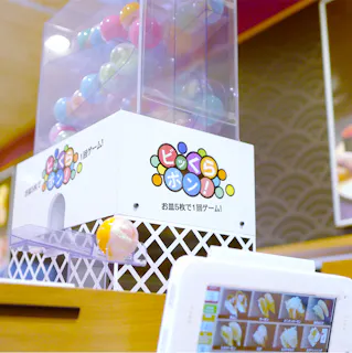 A colorful capsule toy dispenser with Japanese text, releasing a yellow capsule. Capsules are visible inside the machine, and a touchscreen menu displaying sushi items is in the foreground.