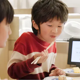 A young child with dark hair wearing a red and white striped sweater uses chopsticks to eat sushi, smiling at a table with another child nearby.