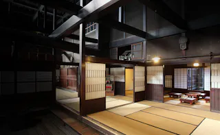 A spacious, traditional Japanese interior with tatami mats, shoji sliding doors, wooden beams, and low tables with floor cushions, softly lit by ceiling lamps. The room has an open, serene, and minimalist atmosphere.