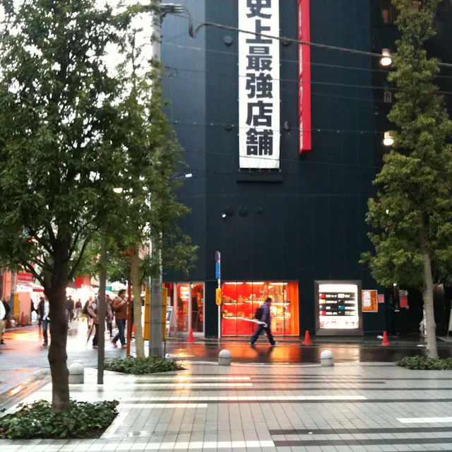 Mandarake Akihabara Complex A city street with people walking, trees lining the sidewalk, and a dark building with a large vertical Japanese sign and bright shop windows on a rainy day.