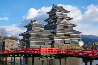 A traditional Japanese castle with black and white wooden walls stands behind a red bridge over a moat, with blue sky and clouds in the background.