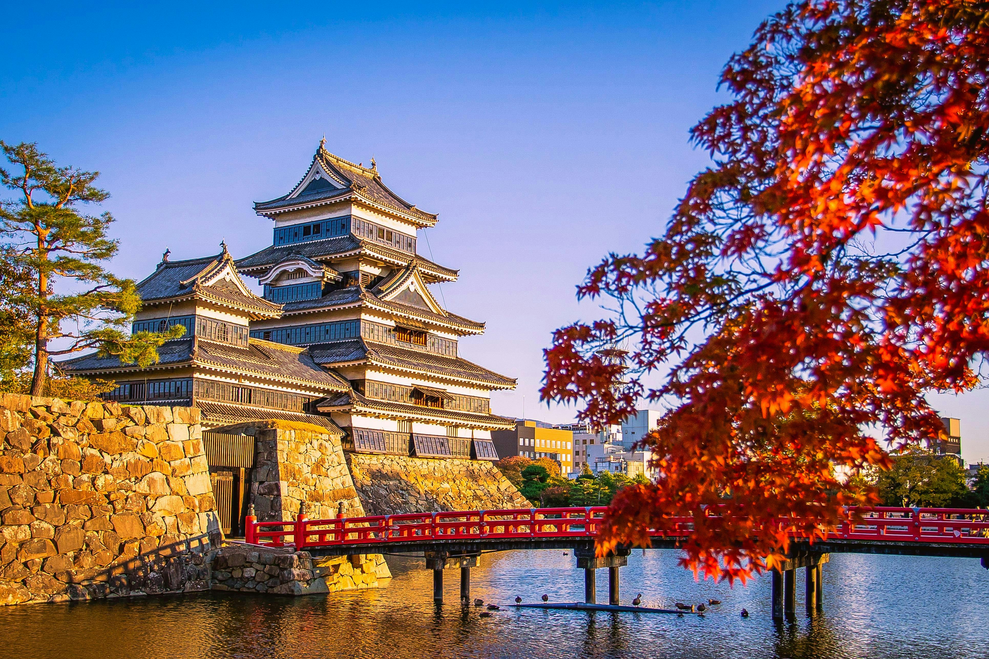 A traditional Japanese castle stands beside a stone wall and reflective moat, with a red wooden bridge and vibrant autumn maple leaves in the foreground under a clear blue sky.