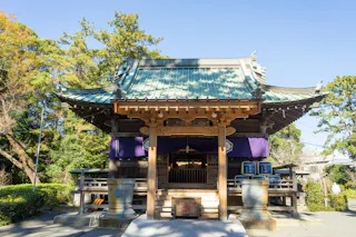 A traditional Japanese Shinto shrine with a green-tiled roof, wooden structure, and purple banners, surrounded by greenery under a clear blue sky.