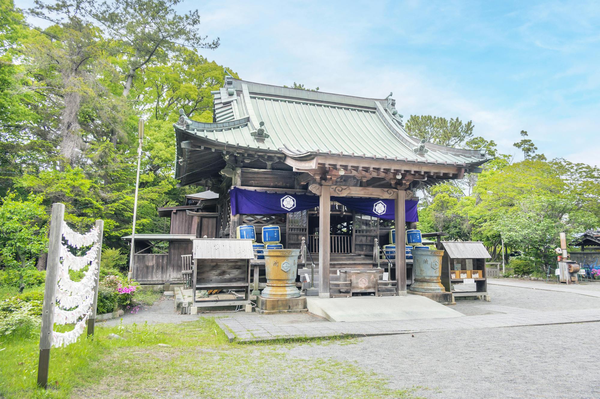 A traditional Japanese shrine with a green tiled roof, wooden beams, and purple banners, surrounded by lush trees and stone lanterns under a bright, clear sky.