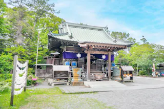 A traditional Japanese shrine with a green tiled roof, wooden beams, and purple banners, surrounded by lush trees and stone lanterns under a bright, clear sky.