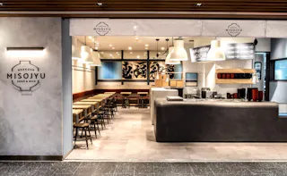 A modern, minimalist restaurant interior with wooden tables and chairs, a sleek gray counter, and pendant lights. The wall has the sign "Asakusa Misojyu Soup & Rice," and the space looks bright and inviting.