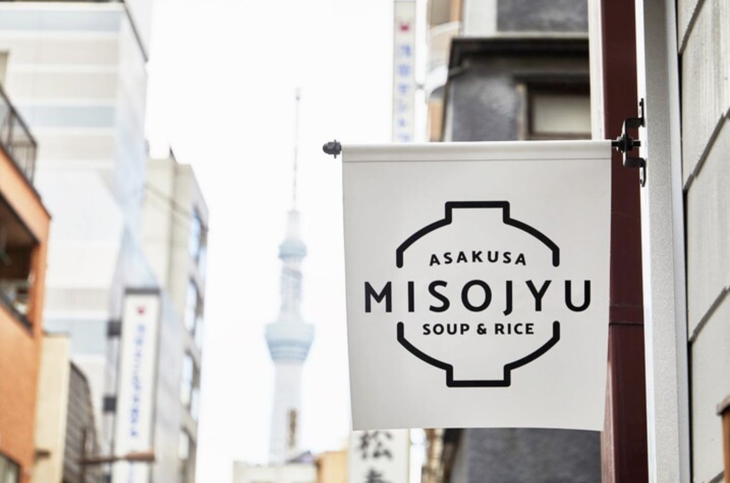 A white hanging sign reads "Asakusa Misojyu Soup & Rice" in black text, with buildings and the blurred Tokyo Skytree visible in the background on a city street.