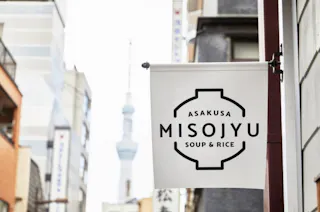 A white hanging sign reads "Asakusa Misojyu Soup & Rice" in black text, with buildings and the blurred Tokyo Skytree visible in the background on a city street.