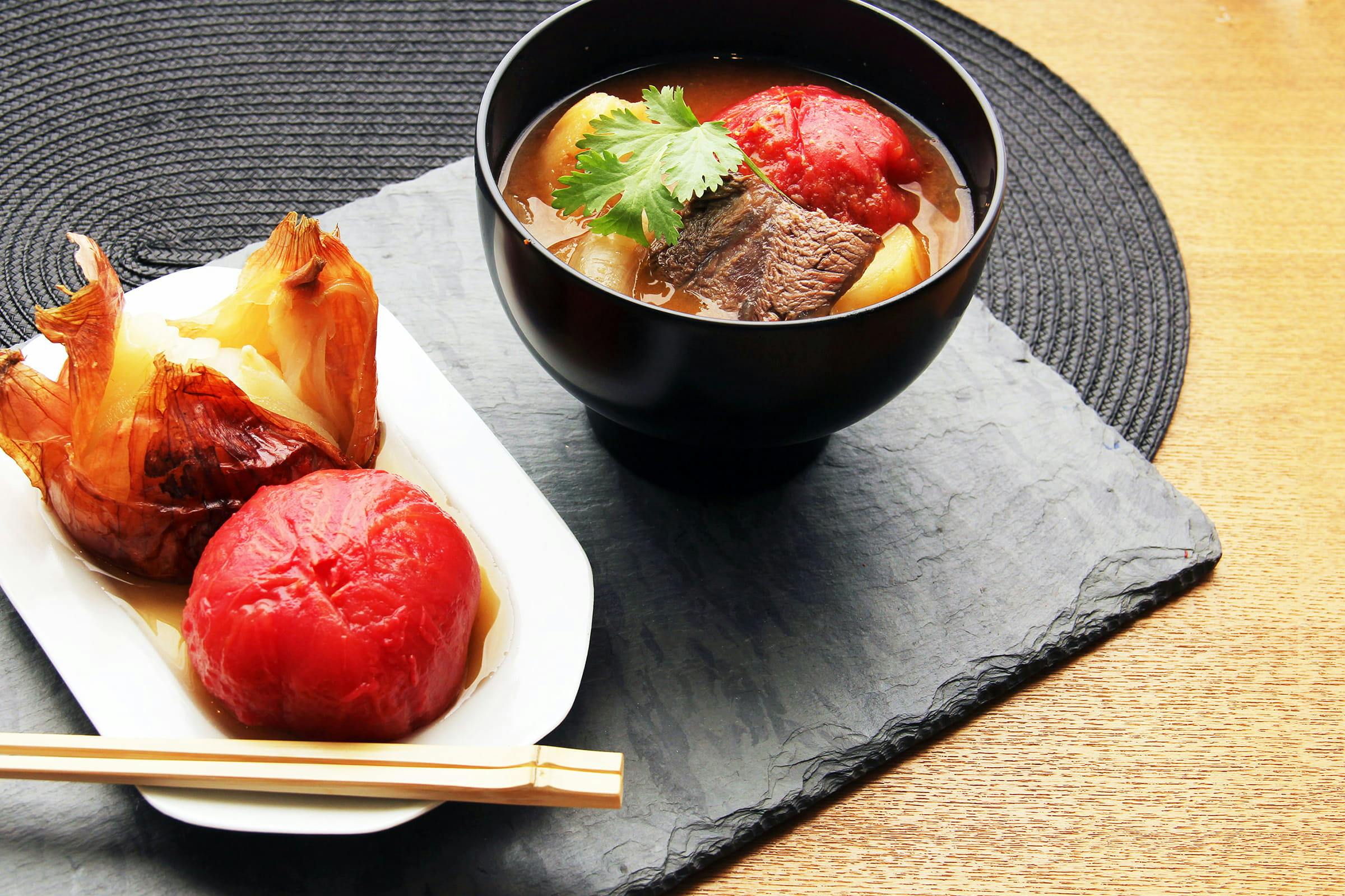 A black bowl of soup with beef, tomato, potato, and cilantro sits on a slate placemat beside a dish with peeled tomato and roasted onion, with chopsticks resting on the plate.