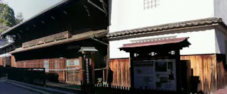 A traditional Japanese wooden building with a dark brown exterior stands next to a white-walled structure. There is an information board and a wooden fence in front, with sunlight casting shadows on the scene.
