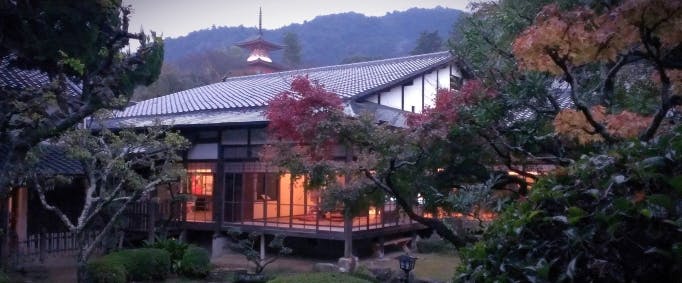 A traditional Japanese house with glowing interior lights sits among trees with autumn foliage, with mountains and a pagoda visible in the background at dusk.