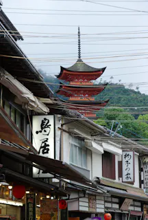 A five-story red pagoda rises above traditional Japanese shops with white signboards and hanging lanterns; mountains and electrical wires are visible in the background.