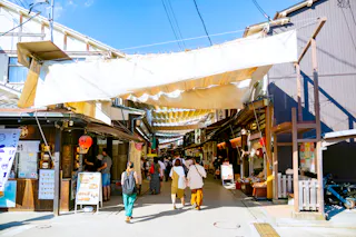 People walk through a bustling outdoor market street lined with shops and restaurants, shaded by large white fabric canopies on a sunny day. Signs, menus, and lanterns are visible along the storefronts.