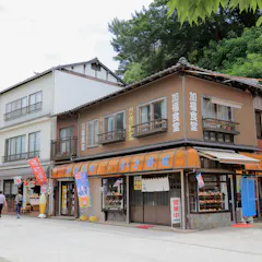 Miyajima Omotesando Shopping Street A street view of traditional Japanese buildings with shops on the ground floor, signage in Japanese, glass display windows, and a person walking in front. Trees and a stone wall are visible in the background.
