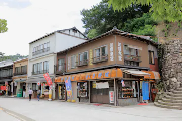 Miyajima Omotesando Shopping Street A street view of traditional Japanese buildings with shops on the ground floor, signage in Japanese, glass display windows, and a person walking in front. Trees and a stone wall are visible in the background.