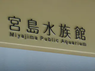 A sign with Japanese characters and English text reading "Miyajima Public Aquarium" on a beige wall.